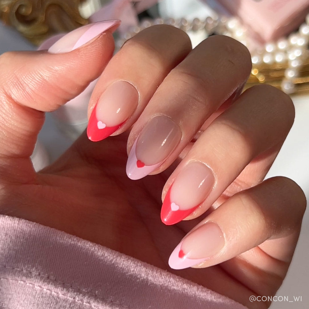 A hand features glamnetics Pure Hearted nails—long almond tips with a pink-to-red French fade and heart accents, all glowing in natural light—an elegant Valentine’s Day look.